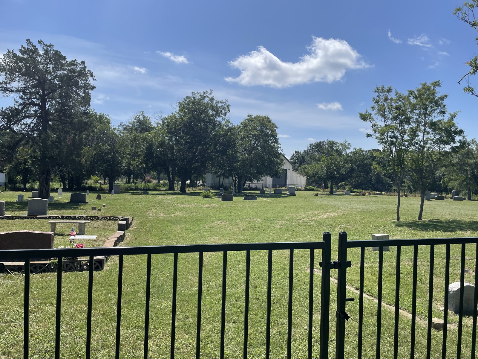 A wide view of the freshly mowed cemetery grounds from the front gate, the schoolhouse and trees beyond.