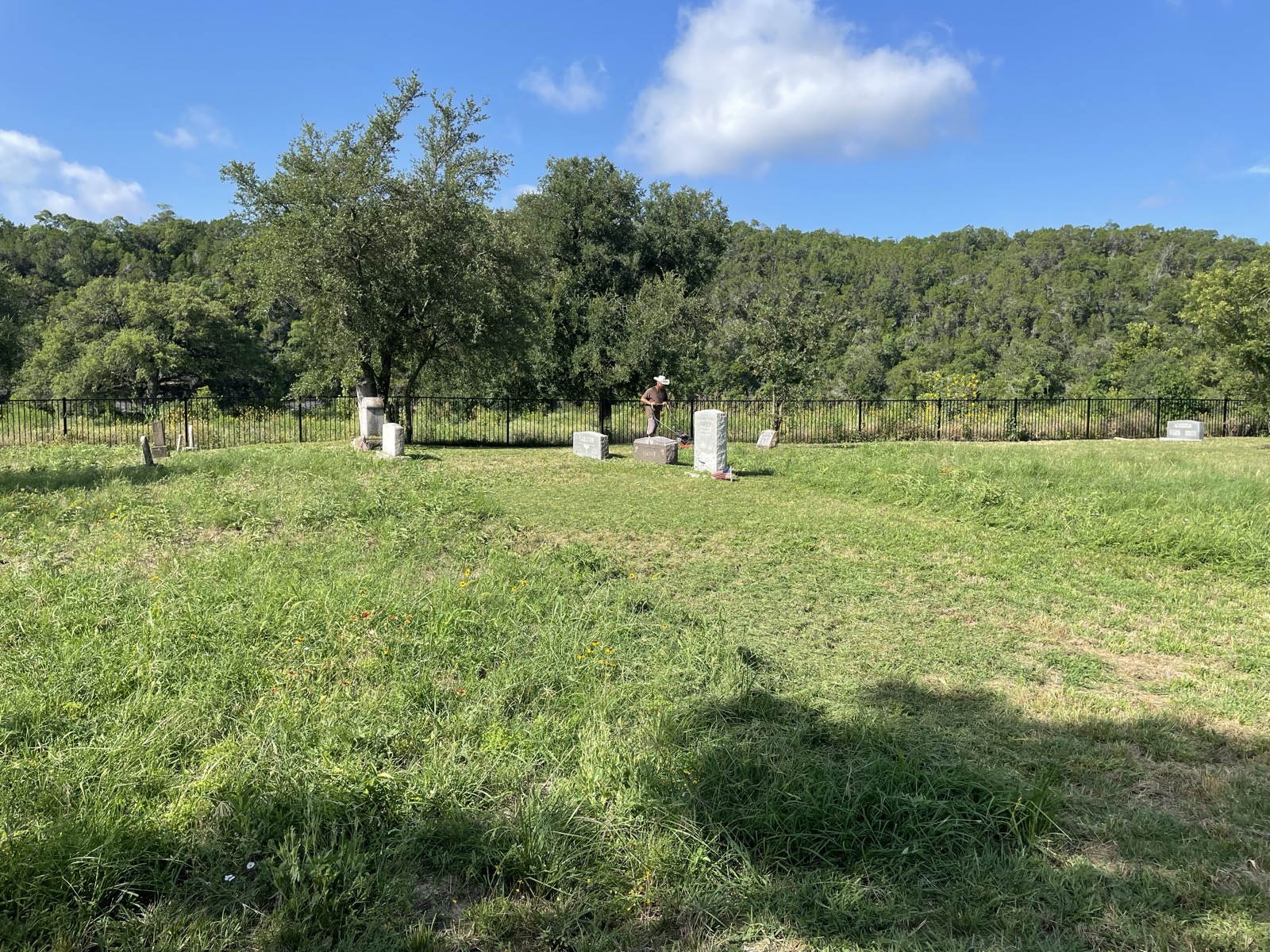 A volunteer trims grass beside weathered grave markers along the tree line.
